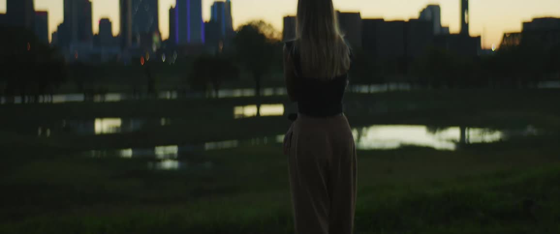 A woman standing in front of a pond with trees and a city skyline in the background
