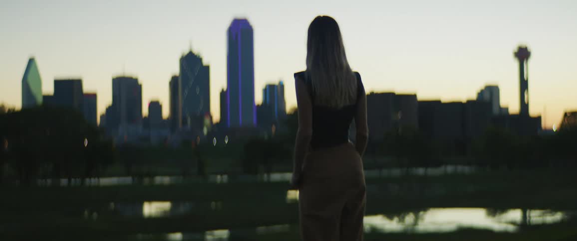 A woman standing next to a pond with trees the city skyline and the sky in the background