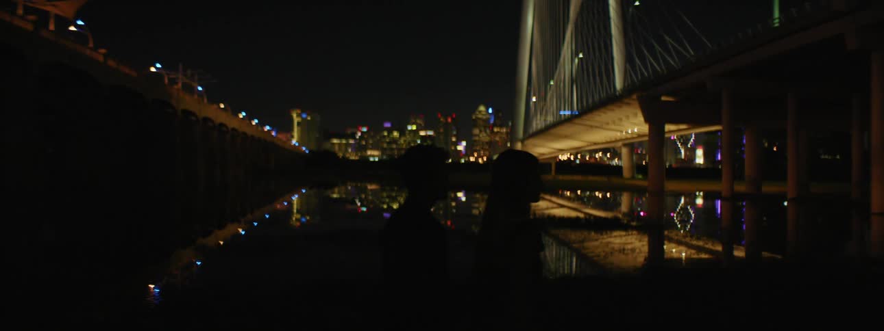 A man and woman walk past a river near a pier across from downtown