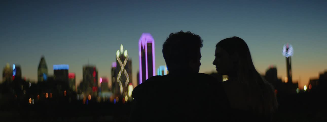 A young man and woman share a kiss while sitting outdoors facing the colorful skyline of a metropolis at sunset