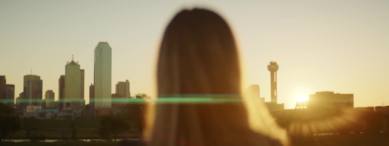 Woman with straight brown hair watches sunset and city skyline