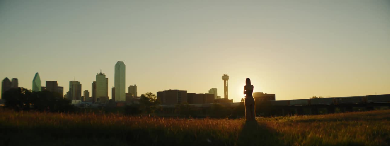 Woman standing in dry grass with tall buildings on the horizon is silhouetted against sunset