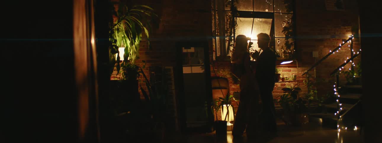 Couple stands close together in dimly lit room near staircase and potted plants