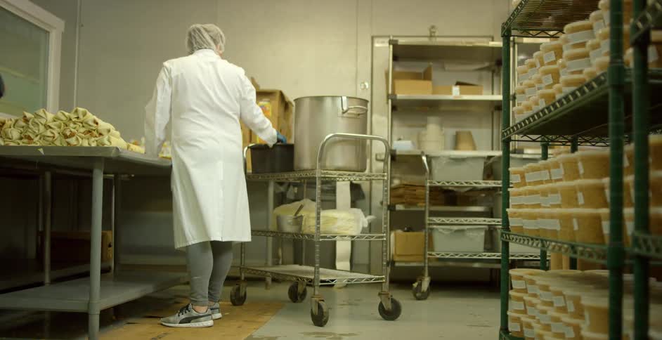 Woman wearing a lab coat and a hair net stirs food in a pot that is sitting on a cart