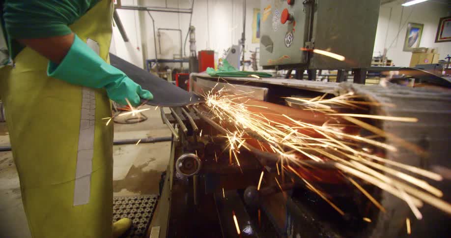 Sparks fly as a man shapes a skateboard on a power belt in a workshop