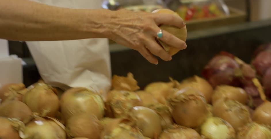 Female reaches for a yellow onion in the produce bin at the grocery store and places it in a white paper bag