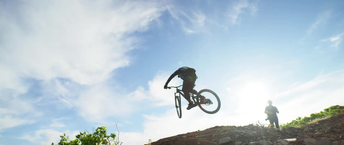A dirt bike rider in a tie dye shirt jumps over a dirt hill and lands on the other side as a friend watches