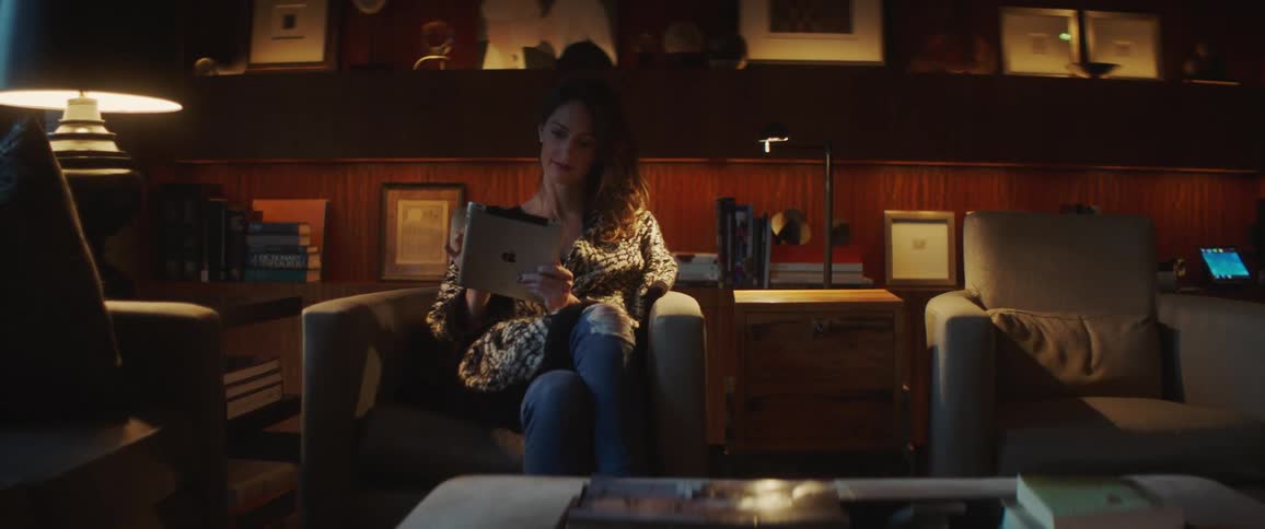 Woman sits in living room reading on tablet near table lamp