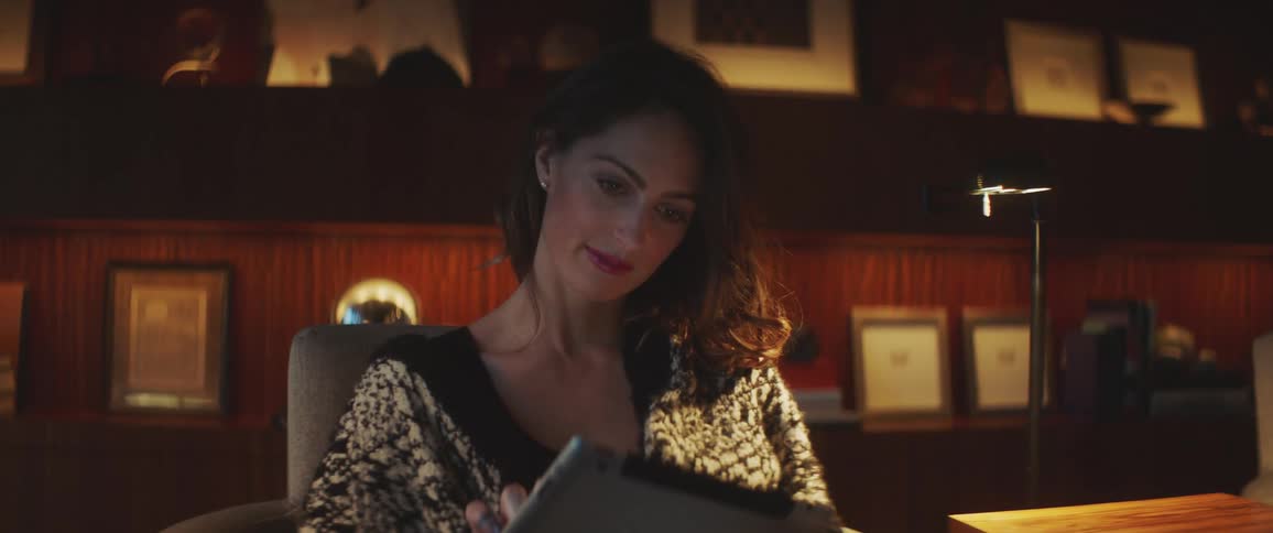A woman sits in a cushioned chair in a dimly lit office and smiles while using a tablet