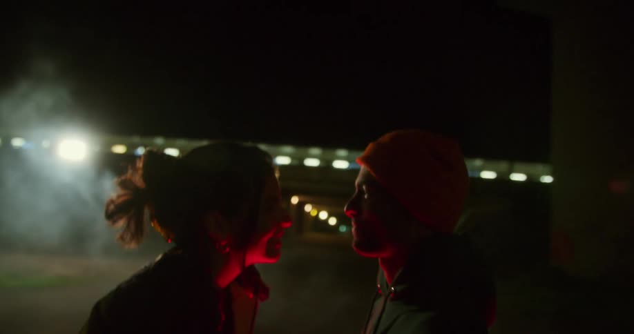 Man and woman face each other at night near car with underpass in distance