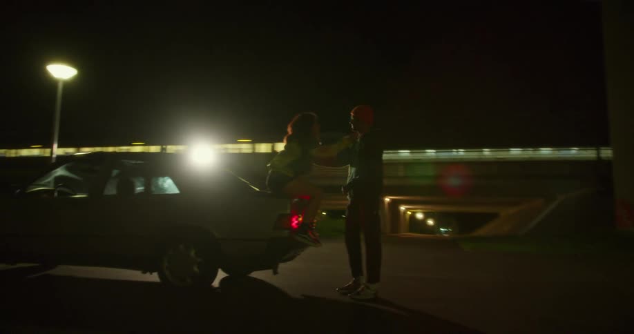 Young couple talk playfully next to a car parked in front of a train station in the city at night