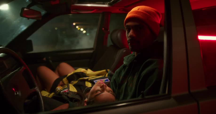A man sits in the drivers seat of a vehicle and looks into the camera as a young woman sitting in the passenger seat rests her head in his lap and speaks to him