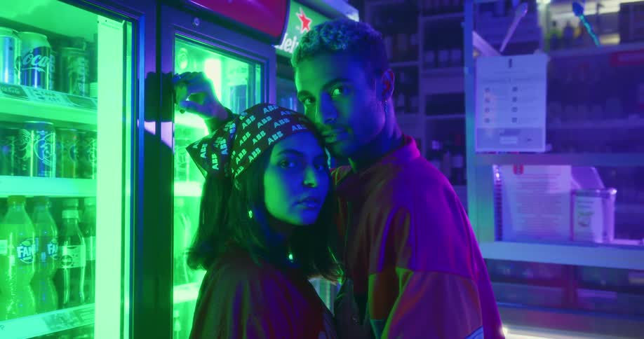 A teenage girl and boy stand in front of a soda machine in a store and look in to one anothers eyes