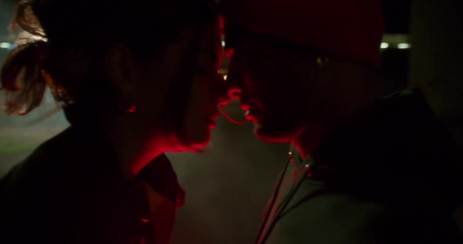 A teenage girl sitting on the trunk of a car pulls a teenage boys close to her as they prepare to kiss