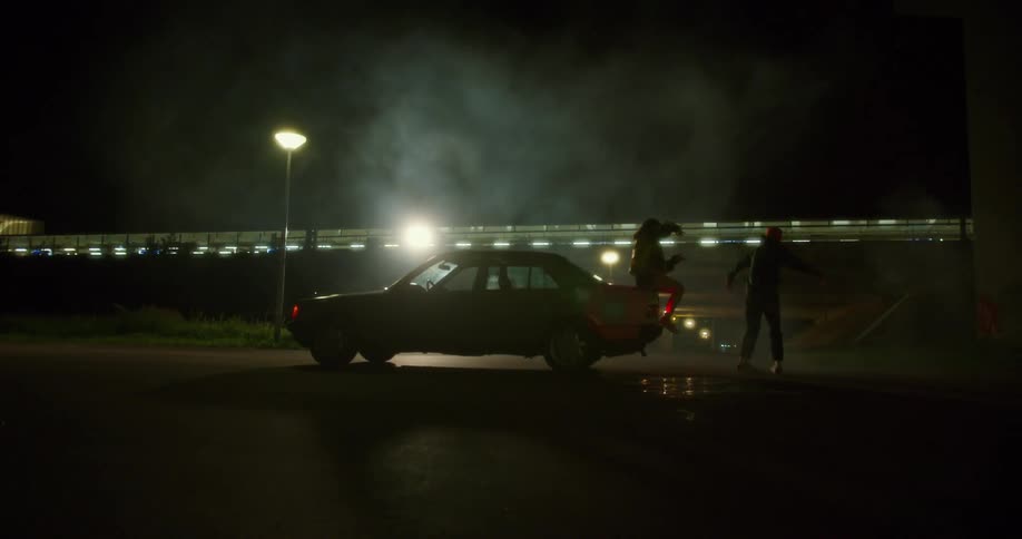 A teenage girl sits on the back of a car and tells a boy to spin around