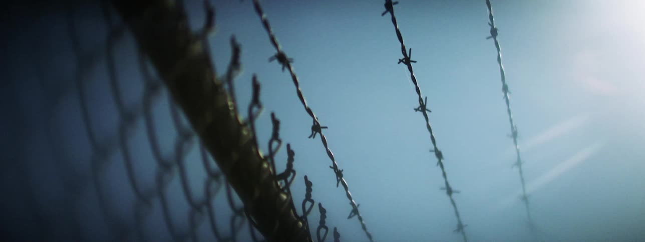 Sunlight shines onto the rows of barbed wire atop a chainlink fence