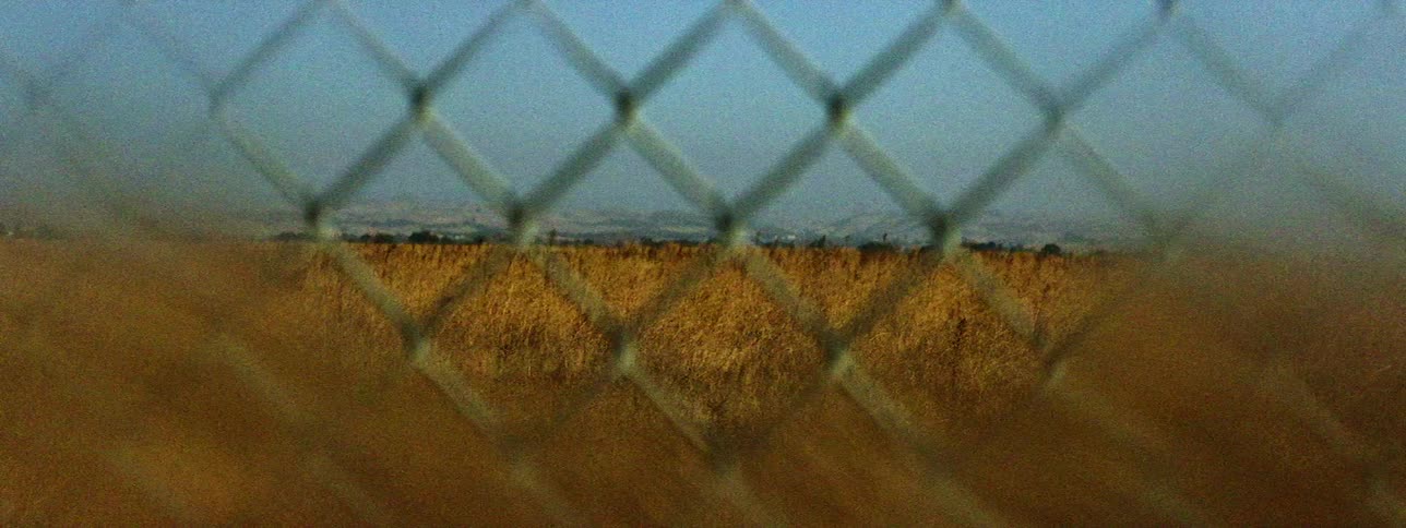 Fog obscured mountains and golden field of grain show through chain link fence