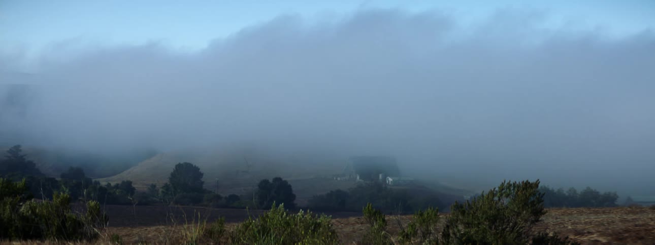 Storm clouds flow over buildings in midst of fields obscuring them and horizon