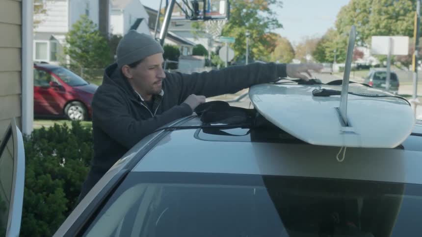 A man secures a surfboard to the top of a car using straps