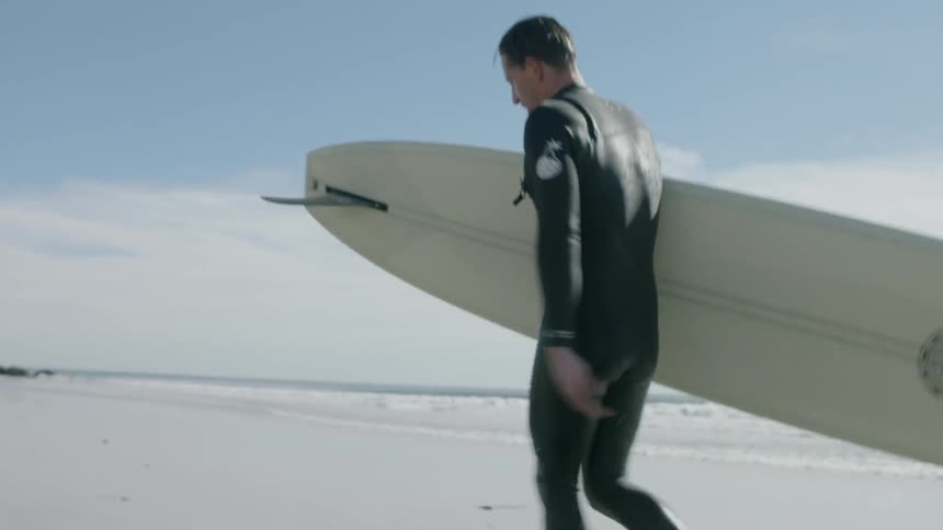A man in a wetsuit walks across the beach carrying a surfboard