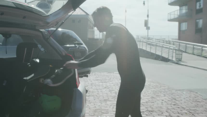 A man in a wetsuit picks up his shoes from the ground and throws them in his suv