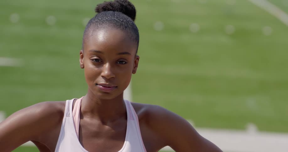 A young black woman standing on an athletics field turns to look into the camera as the sun shines down on her