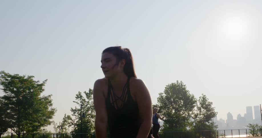 A teenage girl in a public park wipes sweat off of her forehead as another girl jumps rope behind her