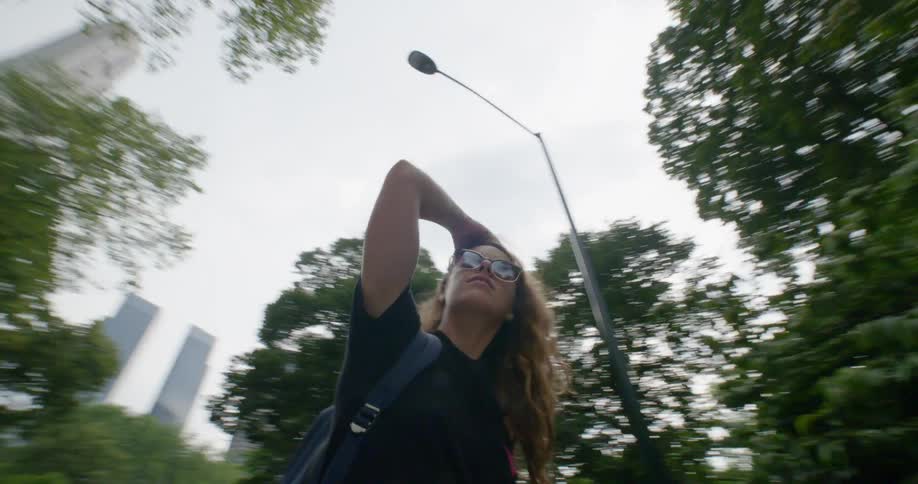 A young woman in sunglasses stands under a street light in a park and gazes around at her surroundings