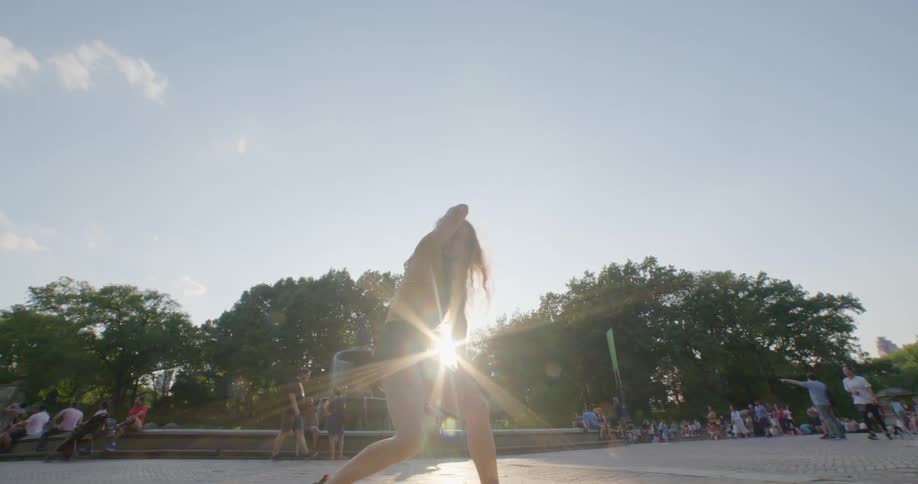 A teenage girl dances in a public park as a large crowd sits and walks around