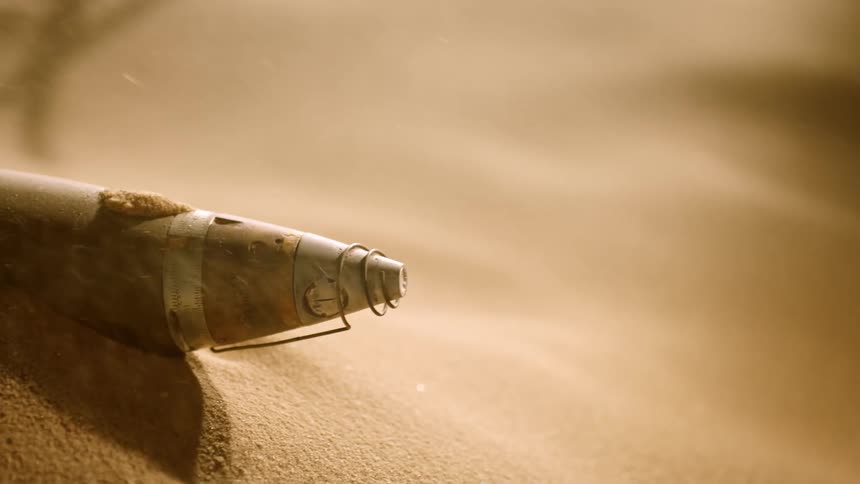 Wooden cylindrical object with metal conical nose wrapped in wire sits half buried among falling sand