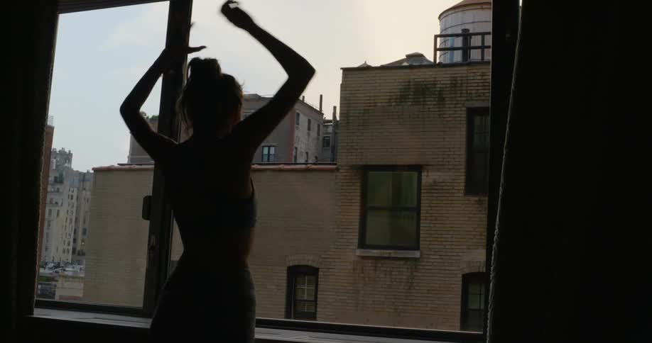 Woman opening window curtains and stretching out is silhouetted against the sky