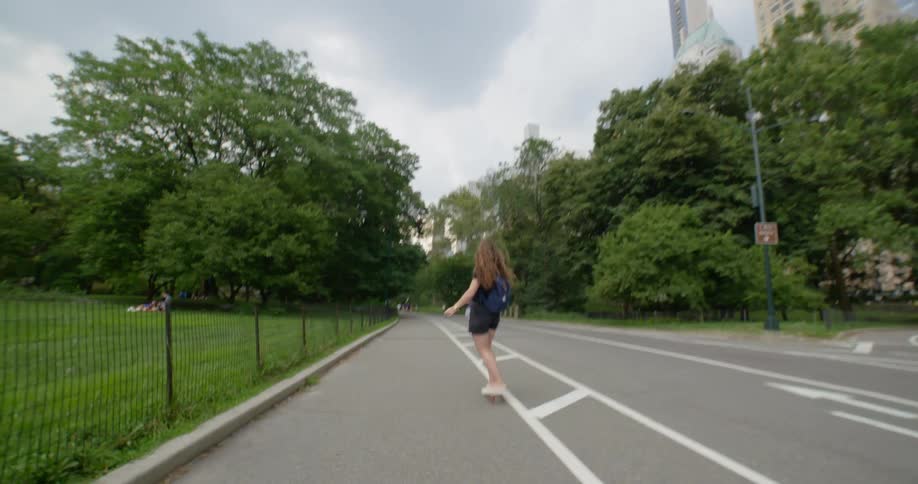 A teenage girl skateboards down a path with other people walking next to a park with people relaxing on the lawn