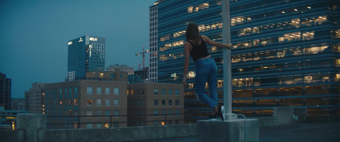 Young woman leisurely hangs from a lamp post on the roof of a parking garage in the city at sunset