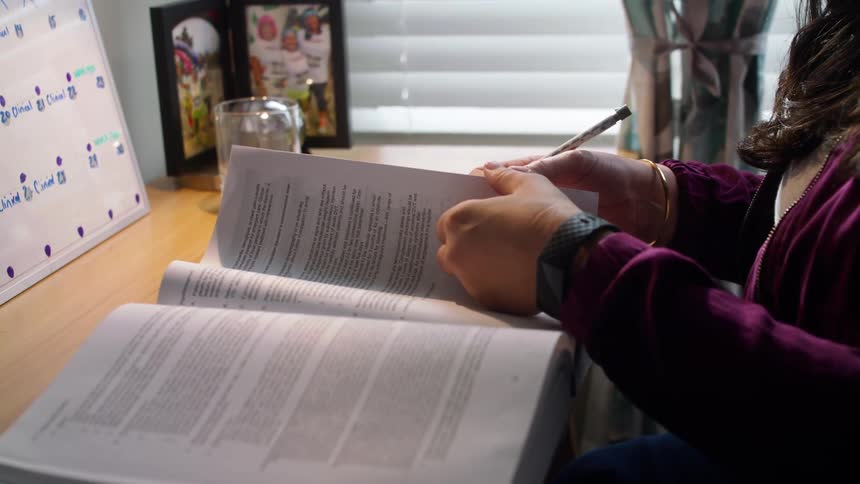 Woman flips through dense textbook at desk holding writing utensil