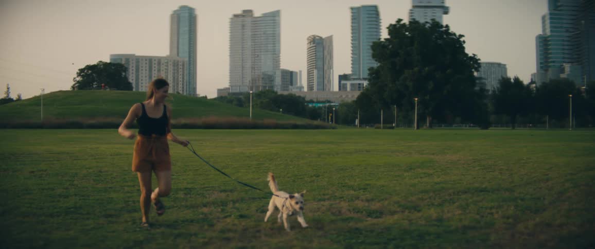 Woman runs with her pet dog on the grass in a park next to the city
