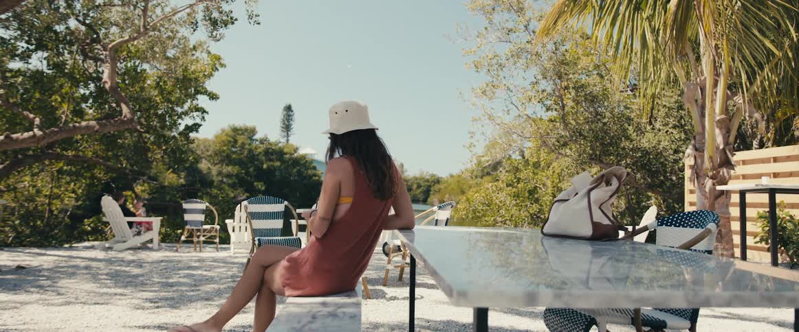A woman with a hat sitting on a bench next to a table with trees in the background