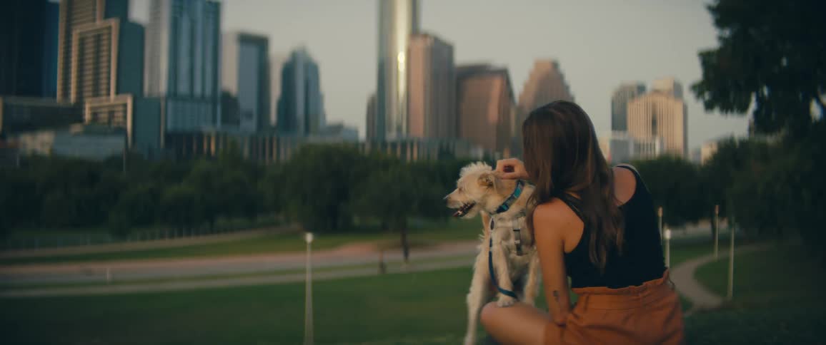 Woman with brown shorts sitting on green grass pets a white dog in public park with tall buildings in the background