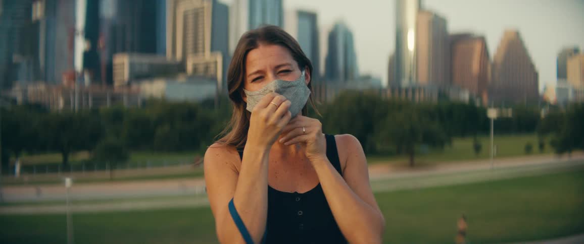 Woman wears a mask in a public park with office buildings in the background at dusk