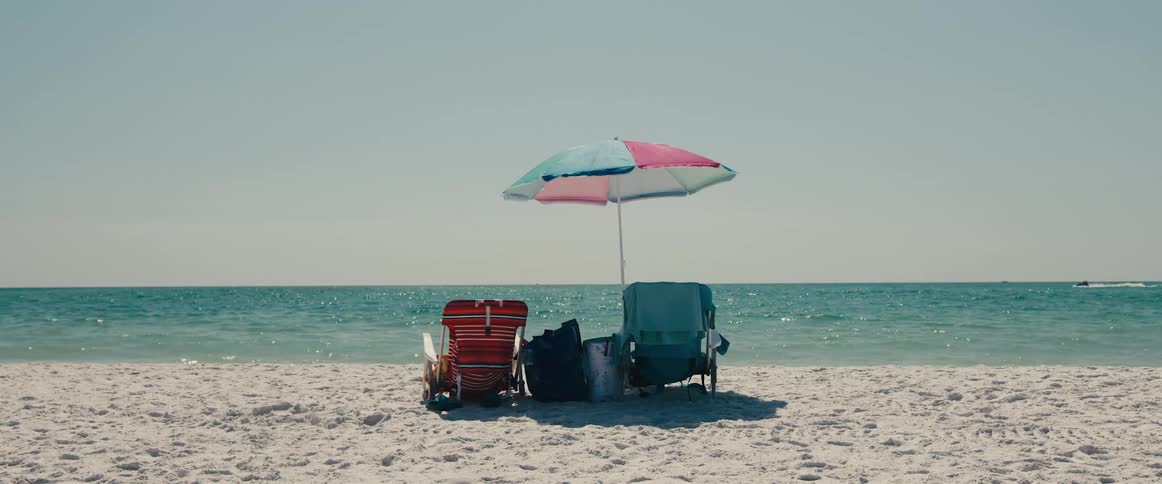 Empty lounge chairs sit under beach umbrella as watercraft speeds across the ocean near the horizon