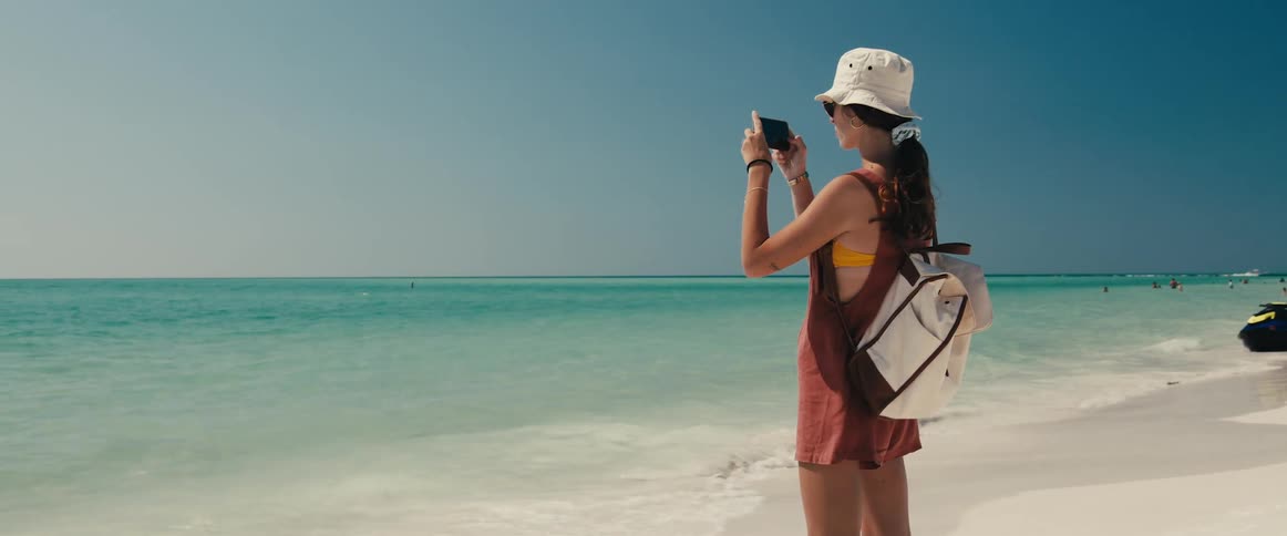 Woman with a white sun hat a backpack and glasses stands on the white sand beach takes pictures of ocean with a smartphone