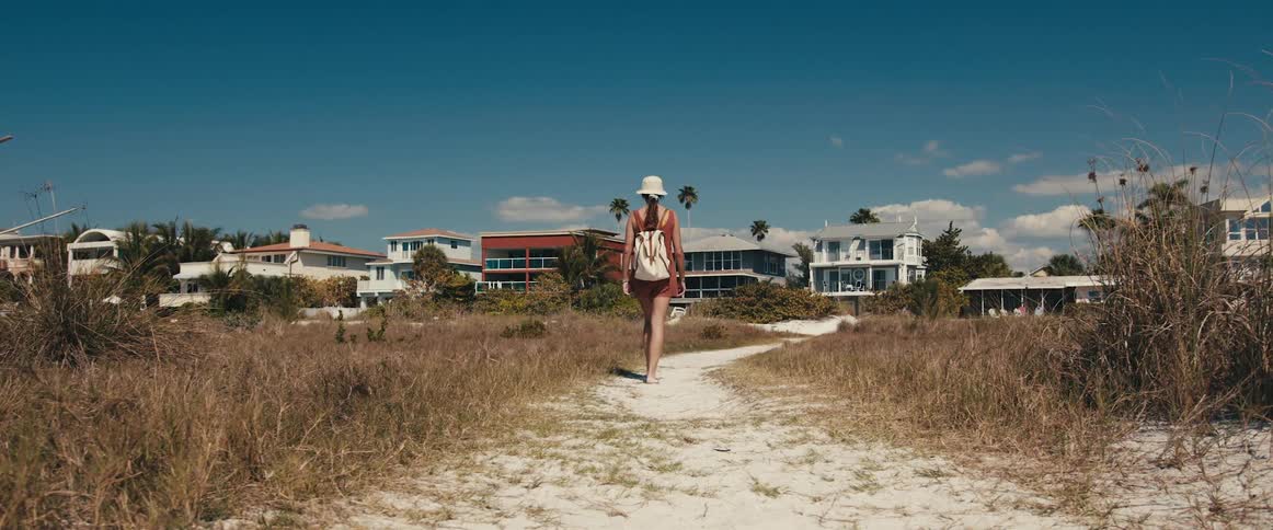 A woman with a backpack and a hat walking on a pathway with houses and trees in the background