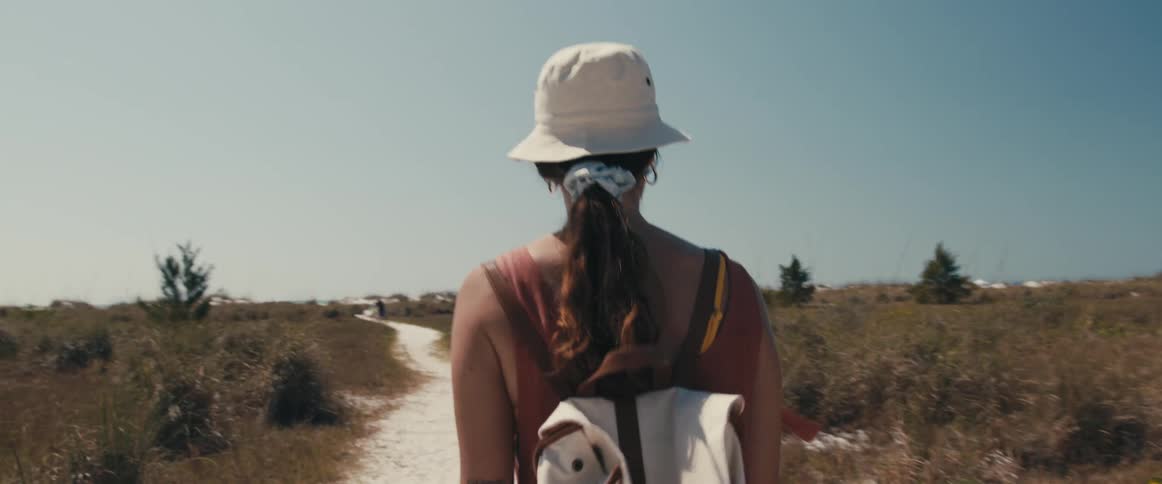 Woman with a brown ponytail a white hat and backpack walks down a dirt road in the open field