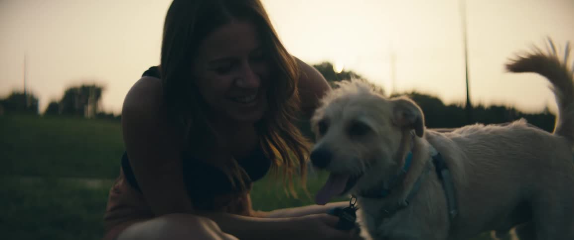 A woman sits in the grass of a public area petting her dog