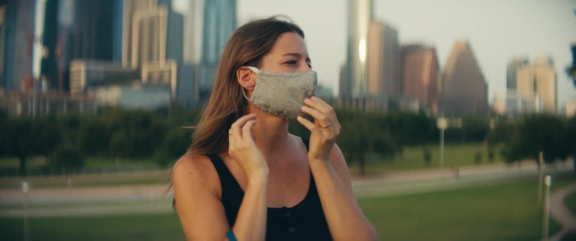 Woman wears a mask in a public park with office buildings at dusk
