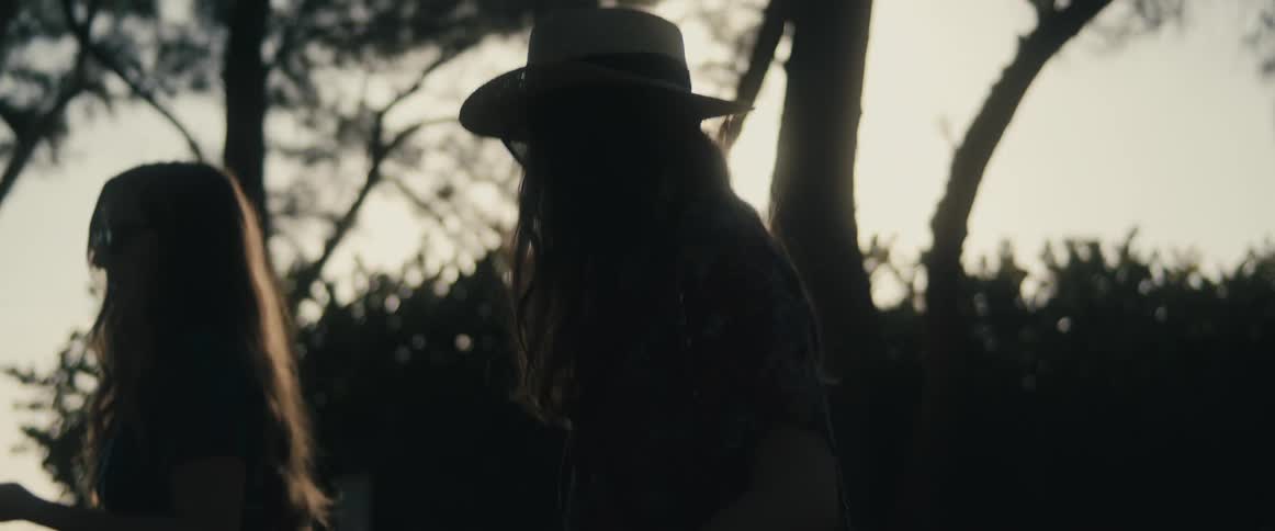 Two young women walk past trees and bushes talking as suns glare turns them into silhouettes