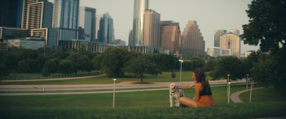 Woman sits in park across from downtown skyscrapers petting a dog