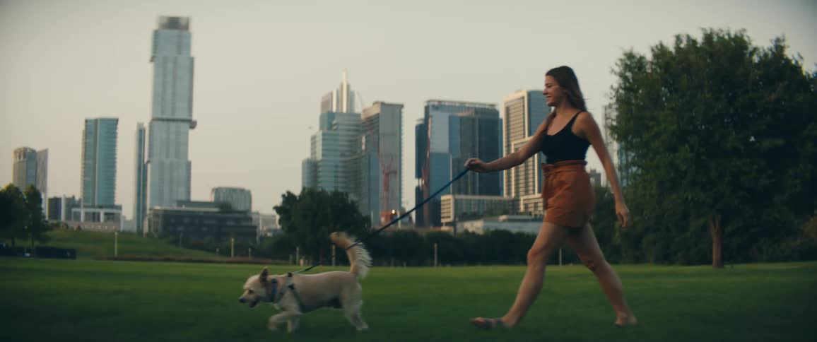 Woman with brown shorts walks a white dog on green grass in public park with tall buildings in the background
