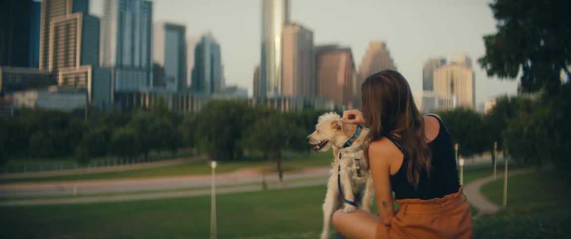 A woman pets her small white dog on the head as they sit in a park across from downtown
