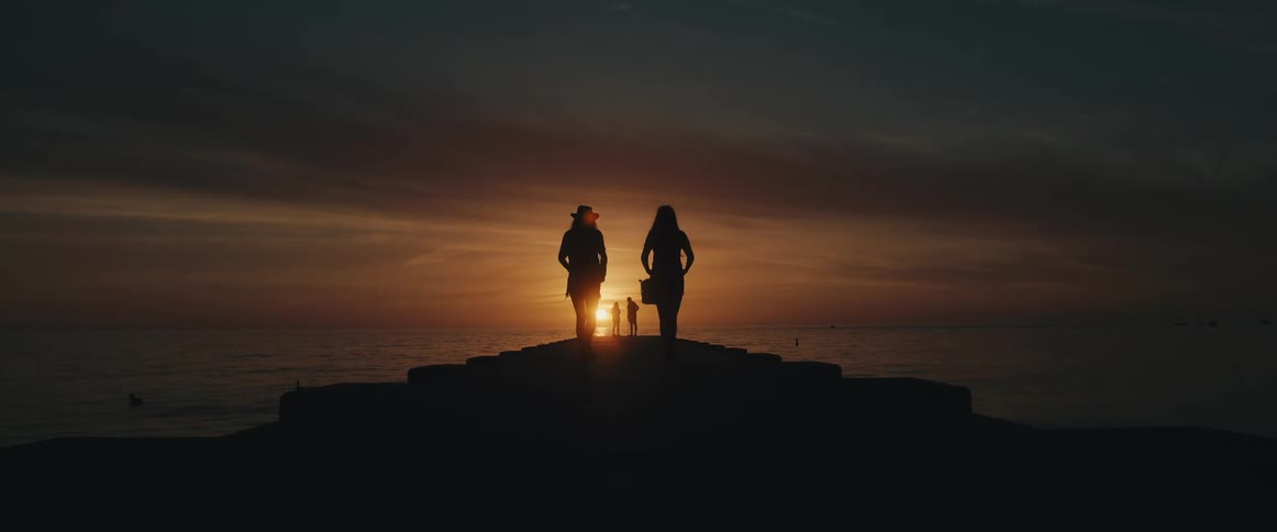 A pair of women walk along a wooden pier towards the ocean as the sun sets behind the horizon