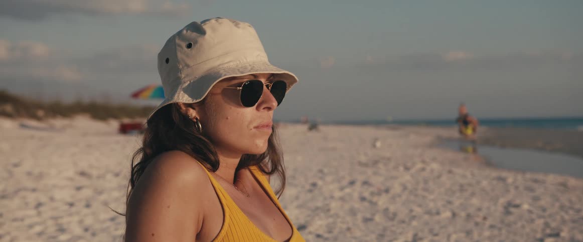 Woman with a sun hat glasses and a yellow bikini top sits on white sand beach next to the sea